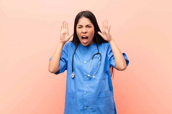 young latin nurse screaming in panic or anger, shocked, terrified or furious, with hands next to head against pink wall