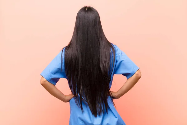 young latin nurse feeling confused or full or doubts and questions, wondering, with hands on hips, rear view against pink wall