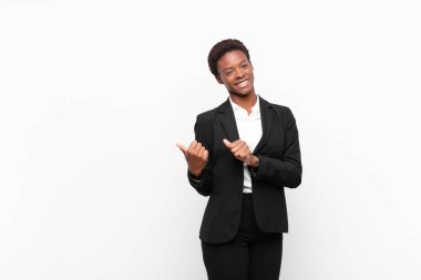 Genç, güzel, siyah bir kadın neşeyle gülümsüyor ve yan taraftaki fotokopi alanını gösteriyor, beyaz duvara karşı mutlu ve tatmin olmuş hissediyor.