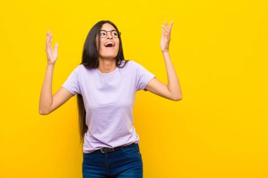 Genç, güzel, Latin bir kadın kendini mutlu, şaşkın, şanslı ve şaşırmış hissediyor. Zaferi kutlarken iki eli de havada, düz duvara karşı.