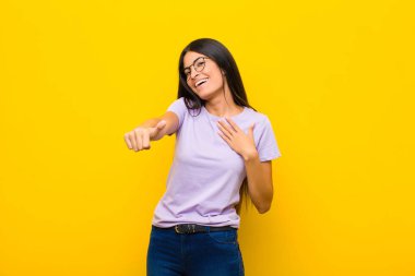 Genç, güzel Latin bir kadın gururlu, tasasız, kendinden emin ve mutlu, parmakları düz duvara değmiş bir şekilde gülümsüyor.