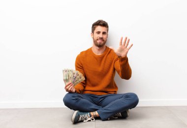 Genç yakışıklı adam gülümsüyor ve arkadaş canlısı görünüyor, 5 ya da 5 numarayı gösteriyor, elinde dolar banknotları tutarak geri sayıyor.