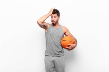 young handsome man raising palm to forehead thinking oops, after making a stupid mistake or remembering, feeling dumb holding a basketball ball.