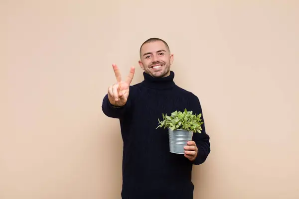 笑顔で幸せそうで気楽でポジティブな若いハンサムな男勝利や平和を手でジェスチャー植物を保持 — ストック写真