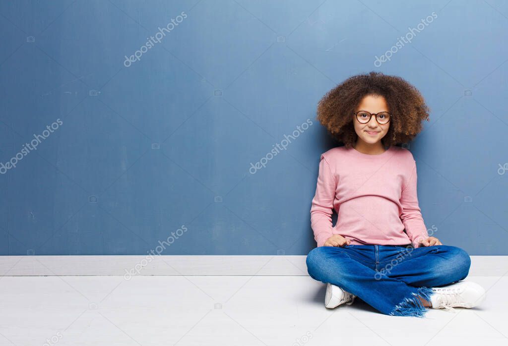 african american little girl smiling positively and confidently, looking satisfied, friendly and happy sitting on the floor