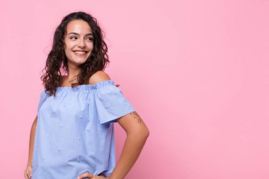 Genç, güzel bir kadın mutlu, neşeli ve kendinden emin görünüyor. Gururla gülümsüyor ve iki elini de pembe duvara yaslamış kalçalarını kolluyor.