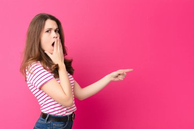 Genç, güzel bir kadın kendini mutlu, şok olmuş ve şaşırmış hissediyor. Ağzını eliyle kapatıyor ve pembe duvara karşı yanal fotokopi alanını işaret ediyor.