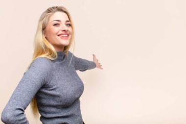Genç ve güzel sarışın kadın mutlu ve neşeli hissediyor, gülümsüyor ve seni selamlıyor, düz duvara karşı dostça bir jestle içeri davet ediyor.