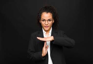 Genç siyahi kadın ciddi, sert, kızgın ve hoşnutsuz görünüyor. Siyah duvara karşı mola işareti yapıyor.
