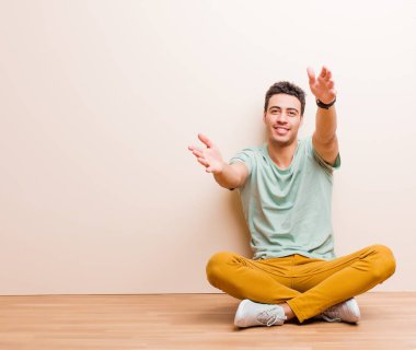 Genç Arap adam neşeyle gülümsüyor sıcak, dostça, sevgi dolu bir kucaklama yapıyor, yerde oturmuş mutlu ve sevimli hissediyor.