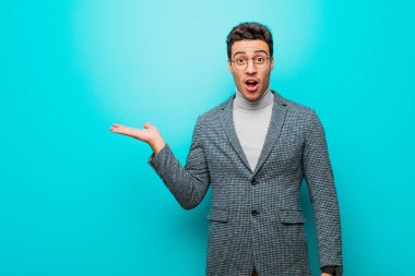 Genç Arap adam şaşırmış ve şoka uğramış görünüyor, çenesi düşmüş, mavi duvara karşı eli açık bir nesneyi tutuyor.