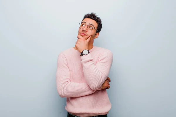 young arabian man feeling thoughtful, wondering or imagining ideas, daydreaming and looking up to copy space against gray wall