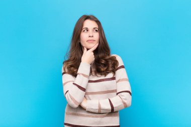 Genç, güzel bir kadın düşünüyor, kuşkulu ve şaşkın hissediyor, farklı seçenekleri var, mavi duvara karşı hangi kararı vereceğini merak ediyor.