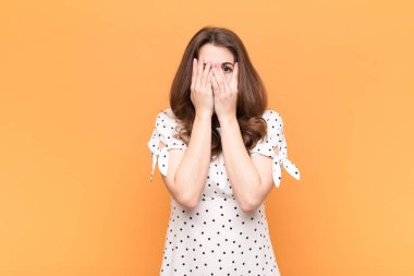 Genç, güzel bir kadın korkmuş ya da utanmış hissediyor. Gözleri turuncu duvara dönük bir şekilde dikizliyor ya da dikizliyor.