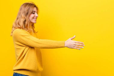 Genç, güzel sarışın kadın gülümsüyor, seni selamlıyor ve başarılı bir anlaşma için el sıkışma teklif ediyor, sarı duvara karşı işbirliği konsepti