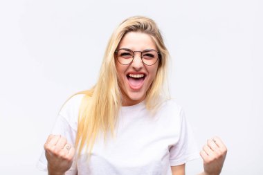 Genç, güzel, sarışın bir kadın muzaffer bir şekilde bağırıyor, gülüyor, mutlu ve heyecanlı hissederken beyaz duvara karşı başarıyı kutluyor.