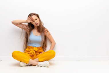 Genç sarışın kadın stresli, yorgun ve hayal kırıklığına uğramış görünüyor, alnından ter akıyor, yerde otururken umutsuz ve bitkin hissediyor.