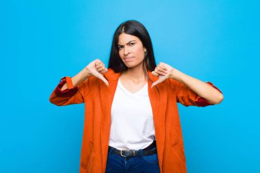 Genç, güzel, Latin bir kadın üzgün, hayal kırıklığına uğramış ya da kızgın görünüyor, anlaşmazlık içinde parmaklarını gösteriyor, düz duvara karşı hayal kırıklığına uğramış hissediyor.