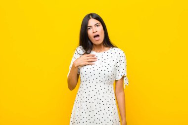 Genç, güzel Latin bir kadın şok olmuş, şaşırmış ve şaşırmış hissediyor, elleri göğsünde ve ağzı açık, kim diyor, ben mi? yassı duvara karşı