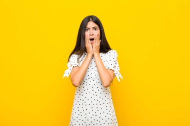 Genç, güzel, Latin bir kadın şok olmuş ve korkmuş hissediyor. Ağzı açık, elleri yanakları düz duvarda.