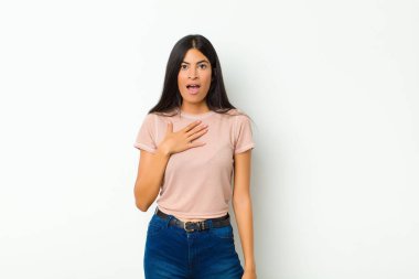 Genç, güzel Latin bir kadın şok olmuş, şaşırmış ve şaşırmış hissediyor, elleri göğsünde ve ağzı açık, kim diyor, ben mi? yassı duvara karşı