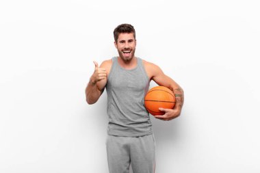 Genç yakışıklı adam gülümsüyor. Mutlu, pozitif, kendinden emin ve başarılı görünüyor. İki başparmağı da basketbol topunu tutuyor..