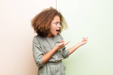 Afro-Amerikan küçük kızı şok olmuş ve şaşırmış hissediyor. Duvara karşı hayret dolu, ağzı açık bir bakışla fotokopi alanını işaret ediyor.