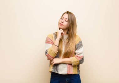 Yoğunlaşmış bakışlı genç sarışın kadın, şüpheli bir ifadeyle, bej duvarın karşısında yukarı ve yana bakıyor