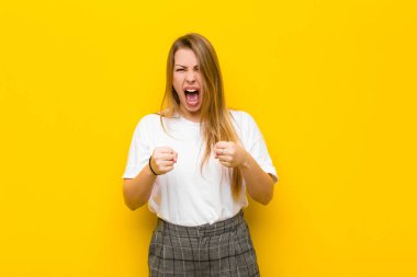 Genç sarışın kadın sinirli sinirli, sinirli bakışlar ve sıkı yumruklarla bağırıyor, turuncu duvara karşı öfkeli hissediyor.