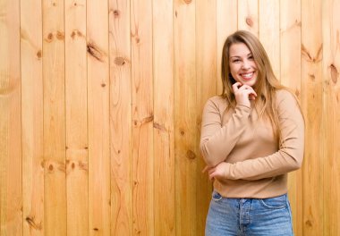 Genç sarışın kadın mutlu görünüyor ve gülümsüyor. Elleri çenesinde, merak ediyor ya da soru soruyor. Seçenekleri karşılaştırıyor.