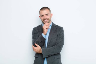 Genç işadamı mutlu görünüyor ve gülümsüyor, merak ediyor ya da soru soruyor, düz duvarla seçenekleri karşılaştırıyor.