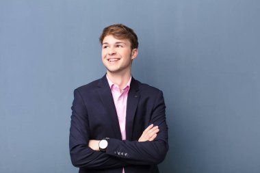 Genç sarışın adam kendini mutlu, gururlu ve umutlu hissediyor, merak ediyor ya da düşünüyor, düz duvara yaslanmış çapraz kollarla fotokopi çekmeyi bekliyor.