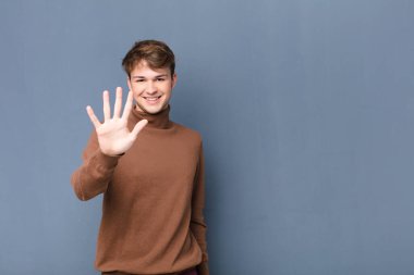 Genç sarışın adam gülümsüyor ve arkadaş canlısı görünüyor. 5 ya da 5 numarayı gösteriyor. Eli önde, geriye doğru sayıyor.