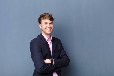 Genç sarışın adam kameraya çapraz kollarıyla gülümsüyor ve mutlu, kendinden emin, tatmin olmuş bir yüz ifadesi var.