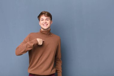 Genç sarışın adam mutlu, gururlu ve şaşırmış görünüyor, neşeli bir şekilde kendini gösteriyor, kendinden emin ve düz duvara karşı soyutlanmış hissediyor.