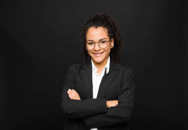 Genç siyahi bir kadın mutlu, gururlu ve tatmin olmuş görünüyor. Kolları siyah duvara kenetlenmiş başarılı bir şekilde gülümsüyor.