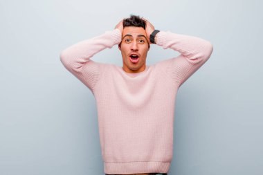 Genç Arap adam heyecanlı ve şaşırmış görünüyor, ağzı açık, iki eli başının üstünde, gri duvara karşı şanslı bir kazanan gibi hissediyor.