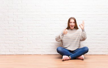 Genç sarışın güzel kız kendini mutlu, şaşkın, tatmin olmuş ve şaşırmış hissediyor, tamam gösteriyor ve parmaklarını kaldırıyor, gülümsüyor yerde oturuyor.