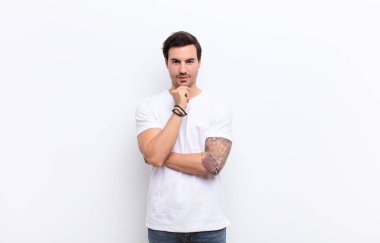 Genç yakışıklı adam mutlu görünüyor ve gülümsüyor, el pençe divan duruyor, merak ediyor ya da soru soruyor, beyaz duvarla seçenekleri karşılaştırıyor.