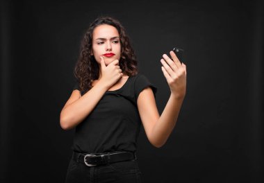 Genç, güzel bir kadın düşünüyor, kuşkulu ve şaşkın hissediyor, farklı seçeneklerle, akıllı telefon tutarak hangi kararı vereceğini merak ediyor.