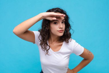 Genç, güzel bir kadın şaşkınlık ve şaşkınlık içinde bakıyor, alnının üstünden uzaklara bakıyor, mavi arka planı izliyor ya da arıyor.