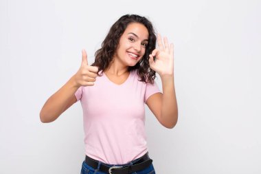 Genç, güzel bir kadın kendini mutlu, şaşkın, tatmin olmuş ve şaşırmış hissediyor. İyi olduğunu gösteriyor ve el hareketlerini gösteriyor. Beyaz duvara gülümsüyor.