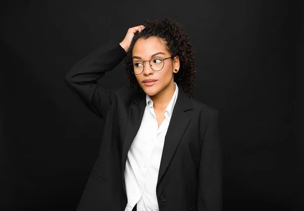 young black woman feeling puzzled and confused, scratching head and looking to the side against black wall