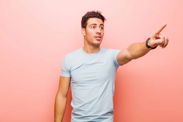 young arabian man feeling shocked and surprised, pointing and looking upwards in awe with amazed, open-mouthed look against pink wall