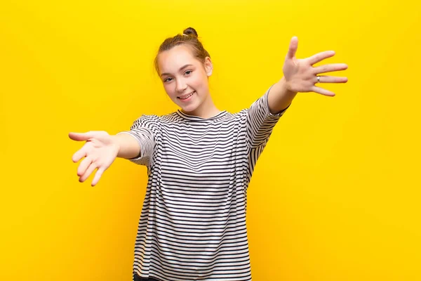 young blonde pretty girl smiling cheerfully giving a warm, friendly, loving welcome hug, feeling happy and adorable against yellow wall
