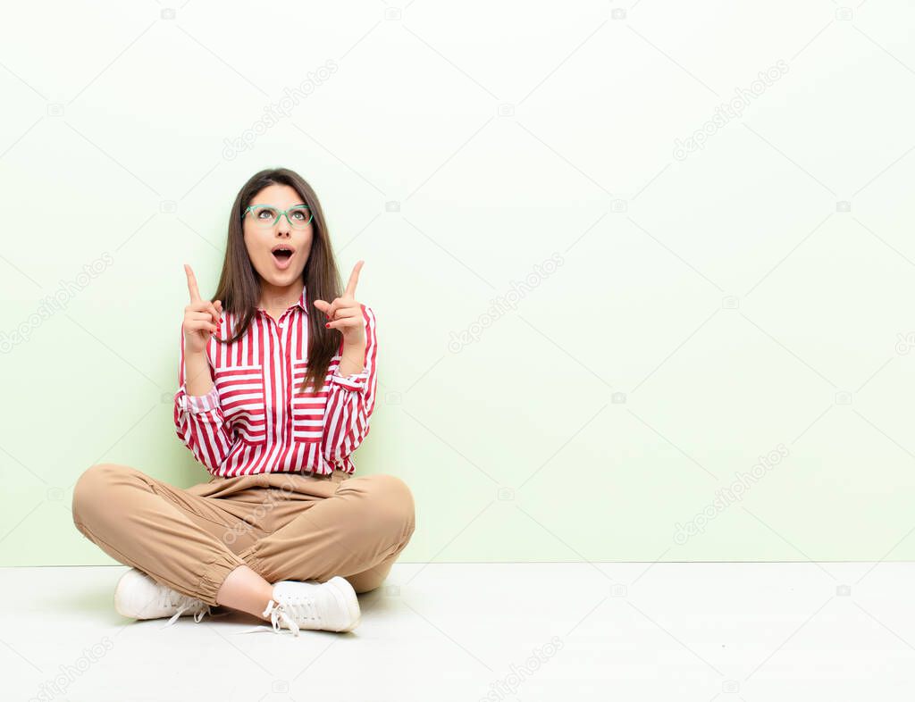 young pretty woman looking shocked, amazed and open mouthed, pointing upwards with both hands to copy space sitting on the floor