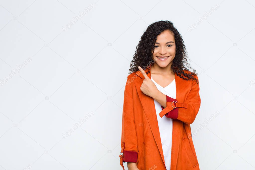 young black woman smiling cheerfully, feeling happy and pointing to the side and upwards, showing object in copy space against white wall