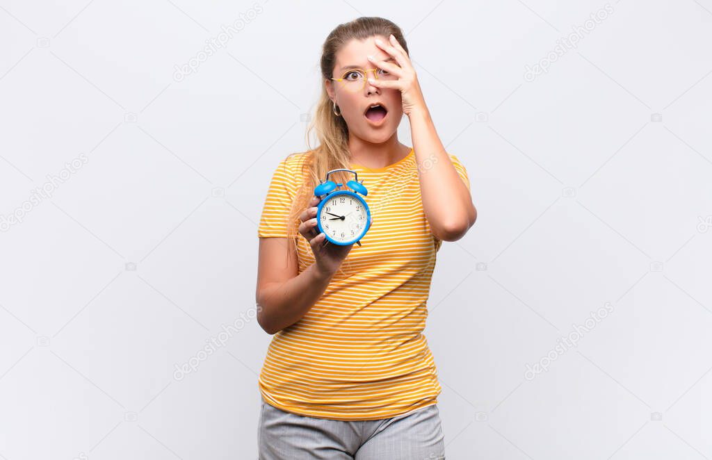 young pretty latin woman looking shocked, scared or terrified, covering face with hand and peeking between fingers with an alarm clock