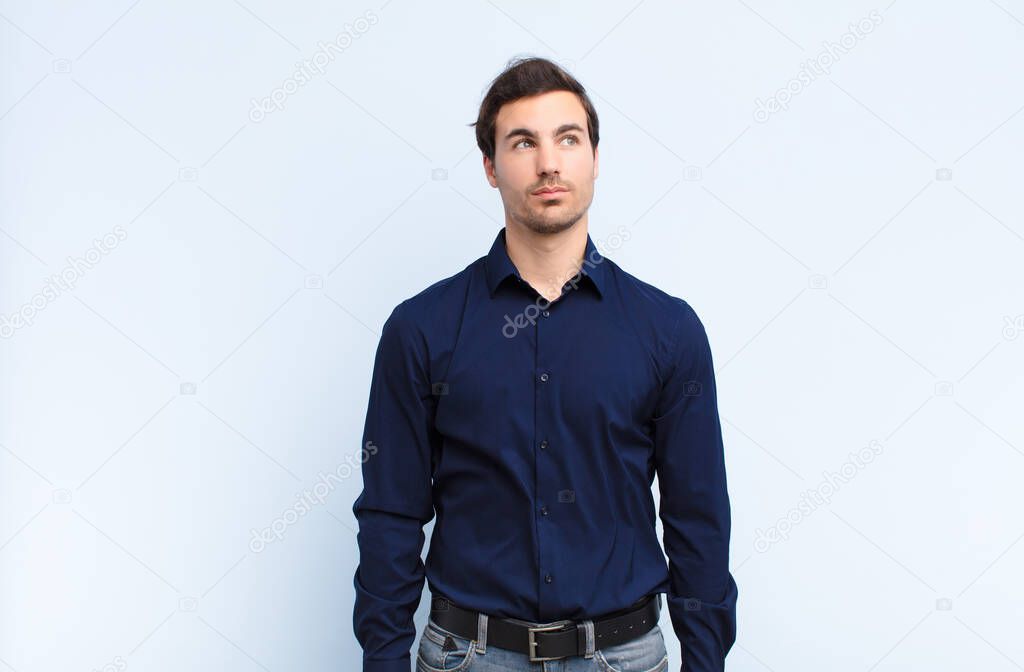 young handsome man with a worried, confused, clueless expression, looking up to copy space, doubting against blue wall