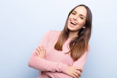 Genç ve güzel bir kadın kollarını kavuşturup mutlu bir şekilde gülüyor. Mavi duvara karşı rahat, pozitif ve tatmin olmuş bir pozla.